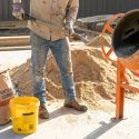 Un ouvrier charge du sable dans une bétonnière orange. À côté, des tas de granulats, un sac de ciment et un seau jaune de 10L.