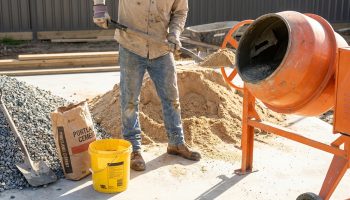 Un ouvrier charge du sable dans une bétonnière orange. À côté, des tas de granulats, un sac de ciment et un seau jaune de 10L.