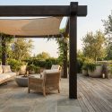 Terrasse avec pergola en bois, mobilier en rotin, plantes en pots en pierre et voile d'ombrage dans un cadre naturel.