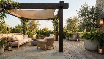 Terrasse avec pergola en bois, mobilier en rotin, plantes en pots en pierre et voile d'ombrage dans un cadre naturel.