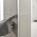 Main gantée grise touchant une baguette de finition métallique insérée entre deux carreaux de carrelage clairs au coin d'un mur.