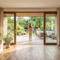 Une femme regarde son jardin depuis un salon lumineux aux menuiseries en bois assorties et parquet en chêne.