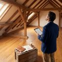 Un expert en costume inspecte un grenier spacieux avec une charpente en bois apparente et une tablette numérique.