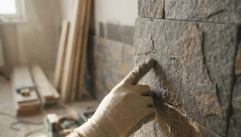 Une main gantée pointe une fissure sur un mur en parement de pierre, dans un environnement de chantier poussiéreux.