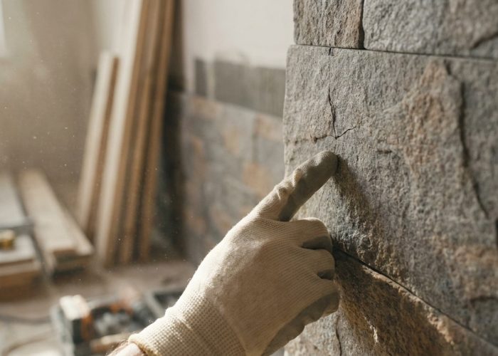 Une main gantée pointe une fissure sur un mur en parement de pierre, dans un environnement de chantier poussiéreux.
