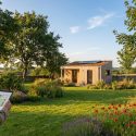 Homme consultant une tablette avec plan de jardin, devant une cabane moderne à toit végétal et panneaux solaires, entourée d'un jardin fleuri.