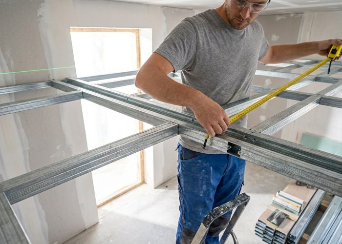 Artisan mesurant une structure métallique pour un plafond autoportant avec un mètre ruban.