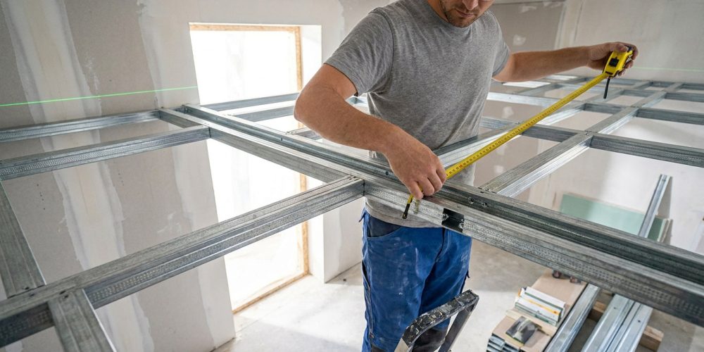 Artisan mesurant une structure métallique pour un plafond autoportant avec un mètre ruban.