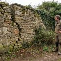 Un homme en tenue de travail inspecte un vieux mur en pierre visiblement penché et fissuré, envahi par la végétation.