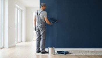 Homme en combinaison grise et casquette peignant un mur en bleu foncé avec un rouleau. Seau et bac à peinture au sol.