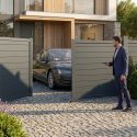 Un homme ouvre un portail battant motorisé gris avec une télécommande devant une maison moderne et une voiture.