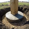 Poteau en bois inséré dans une base en béton coulée dans un trou entouré de graviers pour le drainage.