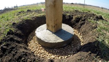 Poteau en bois inséré dans une base en béton coulée dans un trou entouré de graviers pour le drainage.