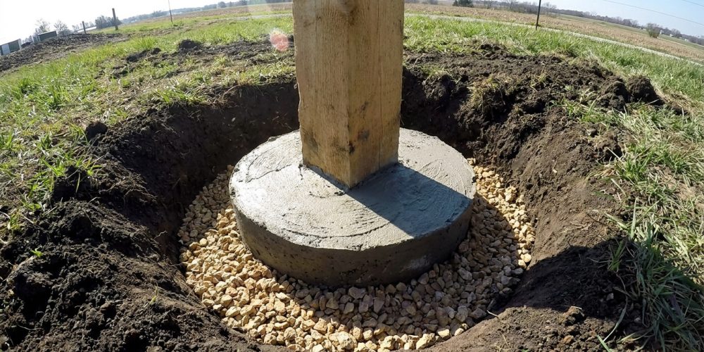 Poteau en bois inséré dans une base en béton coulée dans un trou entouré de graviers pour le drainage.