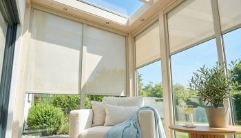 Intérieur de véranda lumineuse avec stores enrouleurs crème baissés, un fauteuil confortable et une plante verte pour le confort estival.
