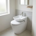 WC blanc avec lavabo intégré compact, robinet chromé, carrelage mural gris clair, fenêtre givrée et serviettes sur étagère.
